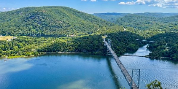 Scenic overlook of the Hudson River with a suspension bridge, rolling green mountains, and forested hills under a bright blue summer sky.