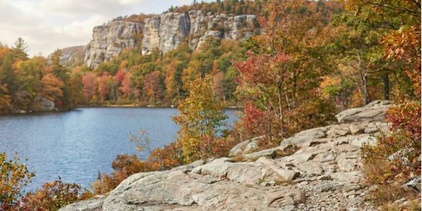 Rocky trail beside a lake in the Hudson Valley during peak fall foliage, with cliffs and colorful trees in the background.