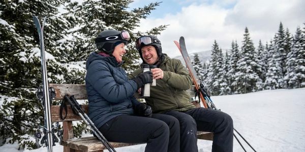 Couple in ski gear sitting on a wooden bench in the snow, sharing a warm drink during a winter day in the mountains